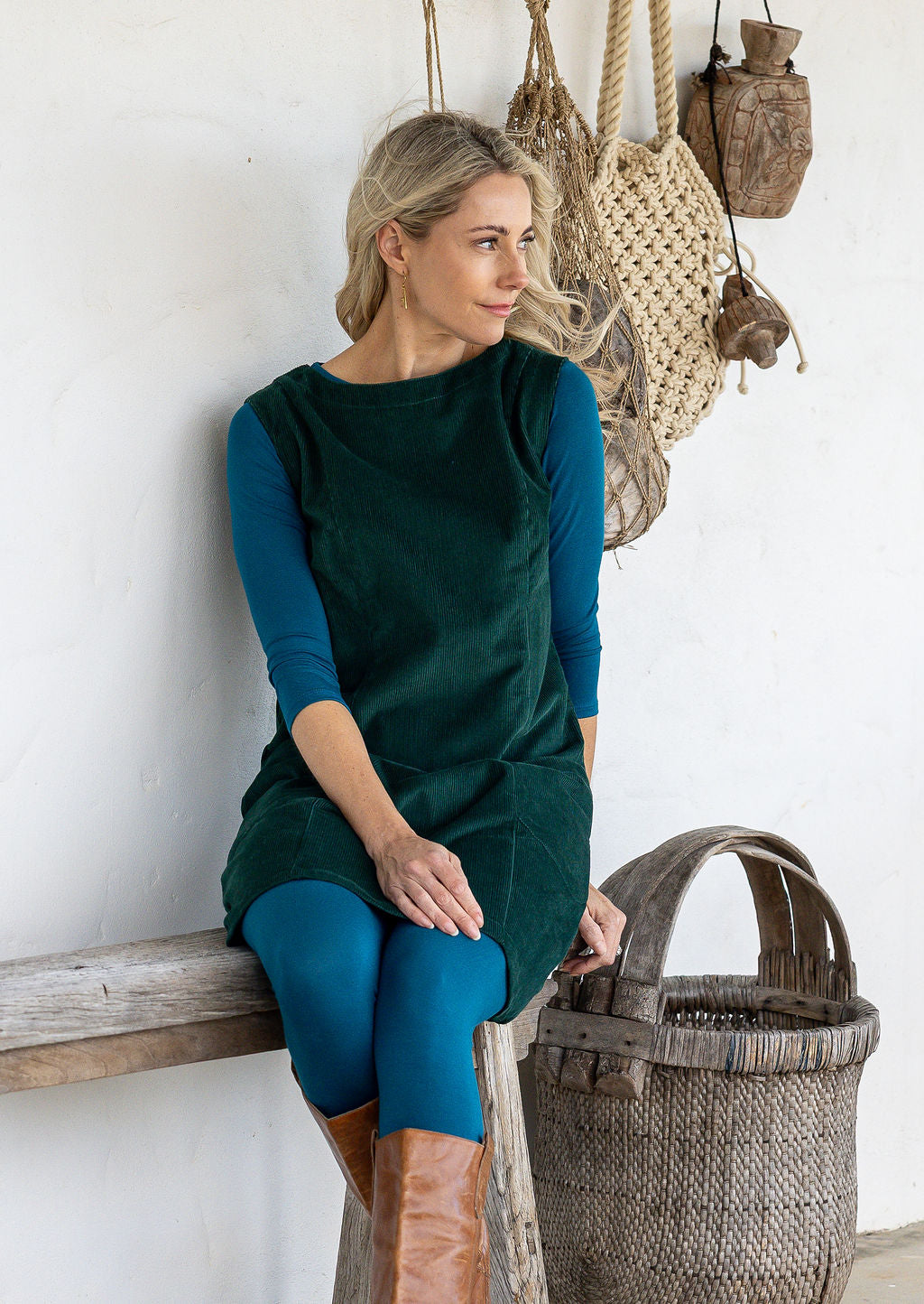 A woman sits on a rustic wooden bench wearing a dark green sleeveless cotton corduroy tunic over a teal long-sleeve top and teal leggings, with tan boots. 
