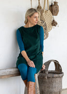 A woman sits on a rustic wooden bench wearing a dark green sleeveless cotton corduroy tunic over a teal long-sleeve top and teal leggings, with tan boots. 