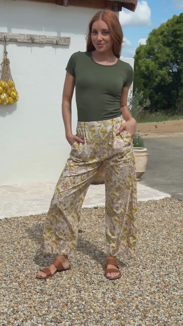 Video of a model wears cream wide-leg trousers with yellow floral print, olive T-shirt, tan sandals, outdoor setting.