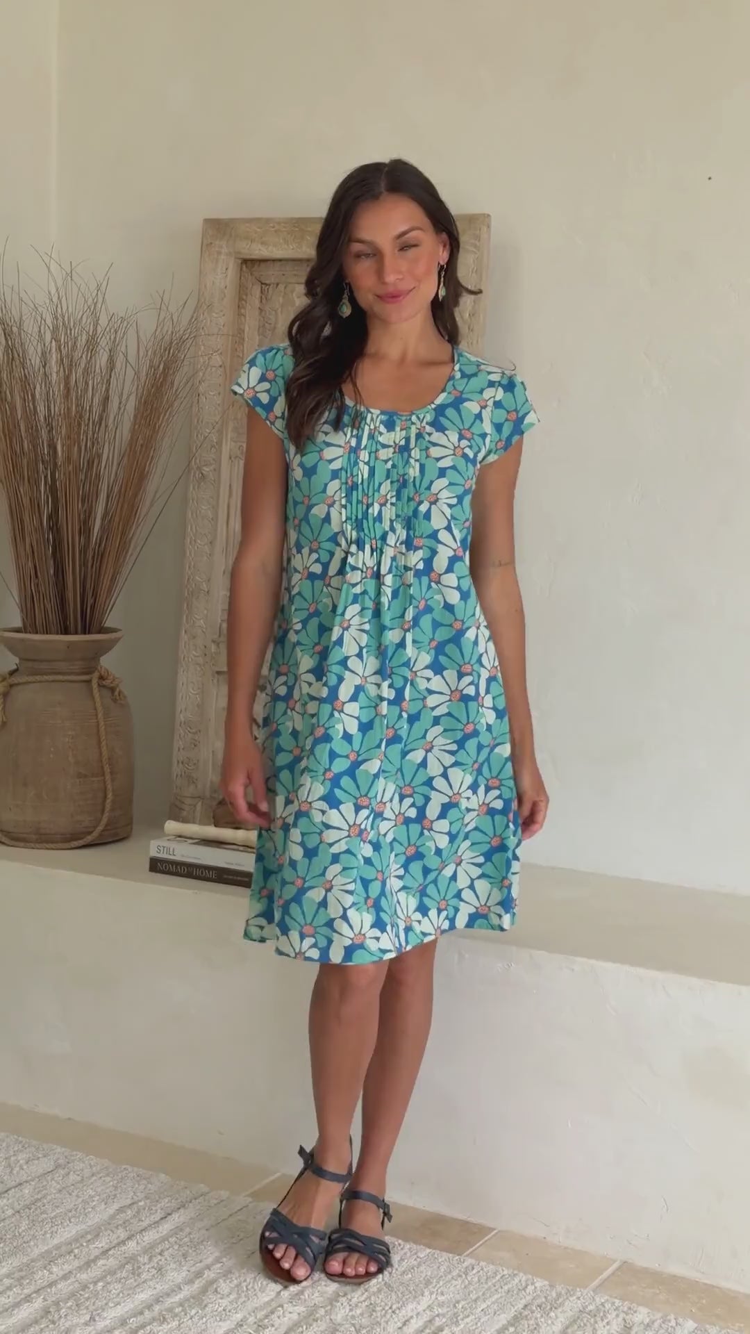 Video of a woman wearing a cap sleeved cotton above knee dress in blue and aqua daisy print, with round neckline and pin-tucks across bodice