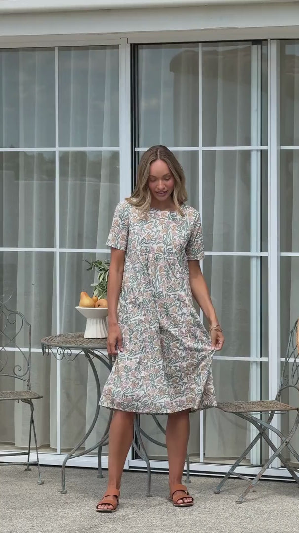 Video of a woman wearing a white based cotton floral short sleeve dress with round neckline and hand in pockets