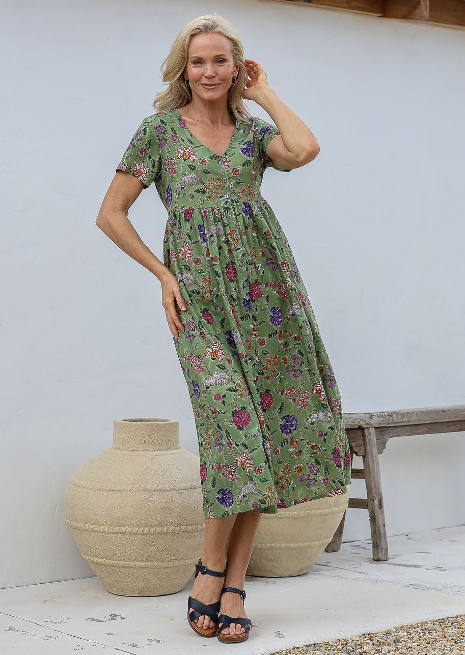 Woman wearing relaxed fit button through dress in moss green based multicoloured floral print with V neckline, pockets and short sleeves.