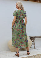 Woman standing in a back pose wearing a 100% cotton relaxed fit dress in moss green with short sleeves, pockets and drawstring waist tie.