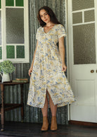 Woman in a white floral cotton dress with buttons down the front standing on a verandah of a rustic cottage