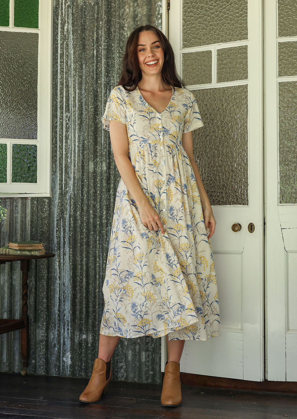 Woman in a white v-neck floral cotton dress standing on a verandah of a rustic cottage smiling 