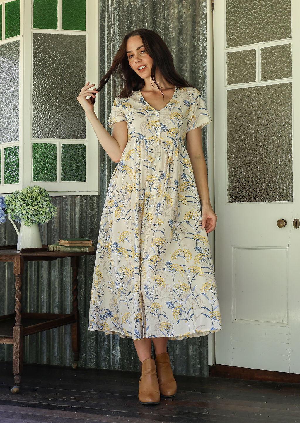 Woman in a white floral cotton dress standing on a verandah of a rustic cottage with her hand in her hair