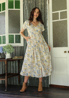 Woman in a white floral cotton dress standing on a verandah of a rustic cottage with her hand in a pocket 