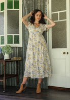 Woman in a white floral cotton dress standing on a verandah of a rustic cottage