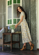 Woman in a white floral cotton dress standing on a verandah of a rustic cottage arranging flowers in a vase