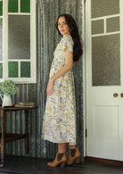 Side view of a Woman in a white floral cotton dress standing on a verandah of a rustic cottage