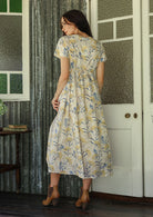 Woman in a white floral cotton dress standing on a verandah of a rustic cottage rear view 