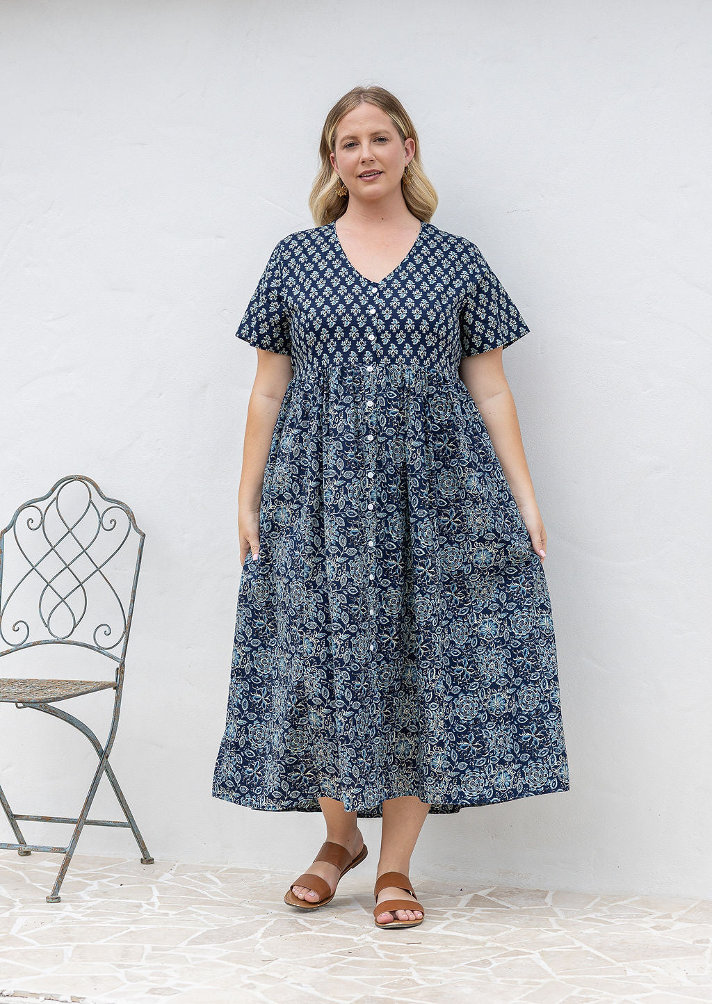 Woman standing in a 100% cotton floral print dress with a V-neckline, short sleeves, pockets, and buttons through to the knee.