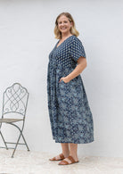 Woman stands sideways wearing a relaxed fit, button through cotton dress in navy blue base floral print with hand in pockets