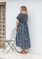 Woman stands sideways wearing a relaxed fit, button through cotton dress in navy blue base floral print with V neckline.