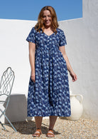A woman wearing a navy blue cotton midi dress with a white botanical print. The dress has short sleeves, a V-neckline, and is button-through. 