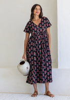 Woman standing in a 100% cotton floral print dress with a V-neckline, short sleeves, pockets, and buttons through to the knee.