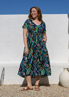 A woman wearing a short-sleeve V-neck cotton midi dress with a colourful floral and peacock print on a dark navy background, with hand in pocket