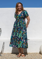 A woman wearing a short-sleeve cotton midi dress with a colourful floral and peacock print on a dark navy background, standing outdoors against a white wall.