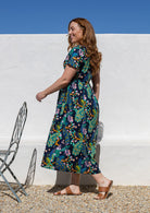 Woman standing side on wearing a short-sleeve cotton midi length dress with a floral and peacock print on a dark navy background, walking outdoors against a white wall under a blue sky.