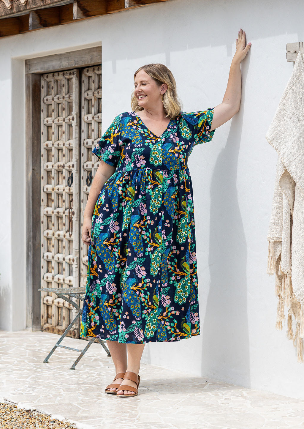 A woman wearing a short-sleeve cotton midi dress with a colourful floral and peacock print on a dark navy background