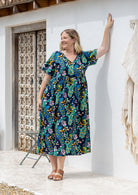 A woman wearing a short-sleeve cotton midi dress with a colourful floral and peacock print on a dark navy background