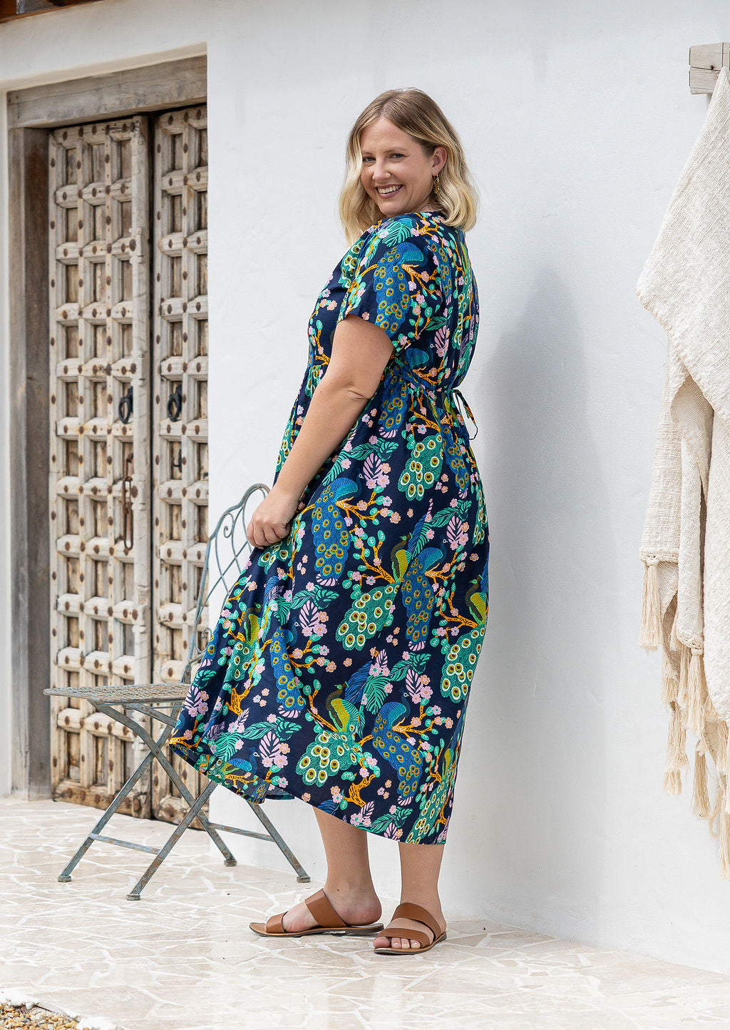 A woman stands sideways wearing a short-sleeve V-neck button through cotton midi length dress with a shoestring ties cinching the waist