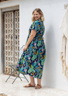 A woman stands sideways wearing a short-sleeve V-neck button through cotton midi length dress with a shoestring ties cinching the waist