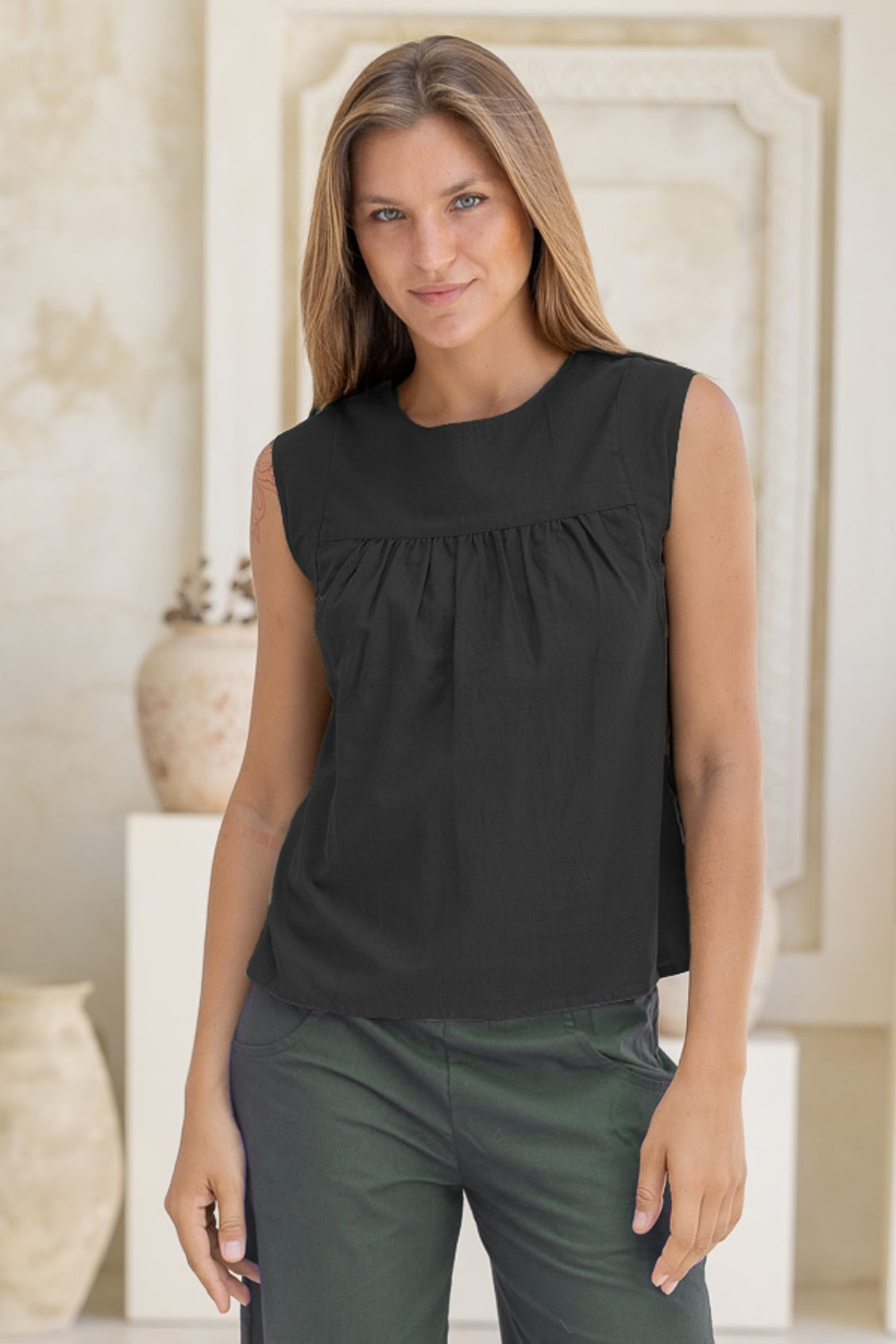 Woman wearing a black sleeveless cotton top  with small gathers across the bust and green pants