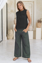 Woman wearing a black sleeveless cotton top with small gather across the bust and green wide-leg pants in a minimalistic room.