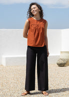 Model stands in plain sleeveless cotton top and black pants against a white wall and a blue sky