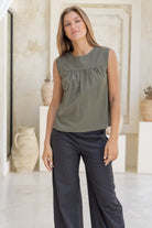 Woman wearing a green sleeveless cotton top and dark wid leg pants in a minimalistic room with decorative vases.