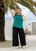 A woman stands outdoors under a palm tree wearing a jade green short-sleeve top and black wide-leg cotton pants, paired with patterned sneakers.