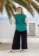A woman viewed from behind wearing a jade green short-sleeve top and black wide-leg cotton pants, standing outdoors on a stone patio under palm tree branches.