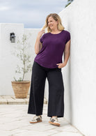 A woman leans against a white wall wearing a short-sleeve purple top and black wide-leg cotton pants with patterned sneakers.