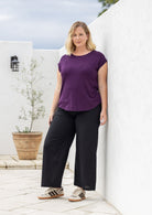 A woman stands outdoors against a white wall wearing a purple short-sleeve top, black wide-leg cotton pants