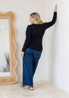 Back view of a woman wearing a black long-sleeve top and cotton navy wide-leg pants, leaning her hand against a white wall.