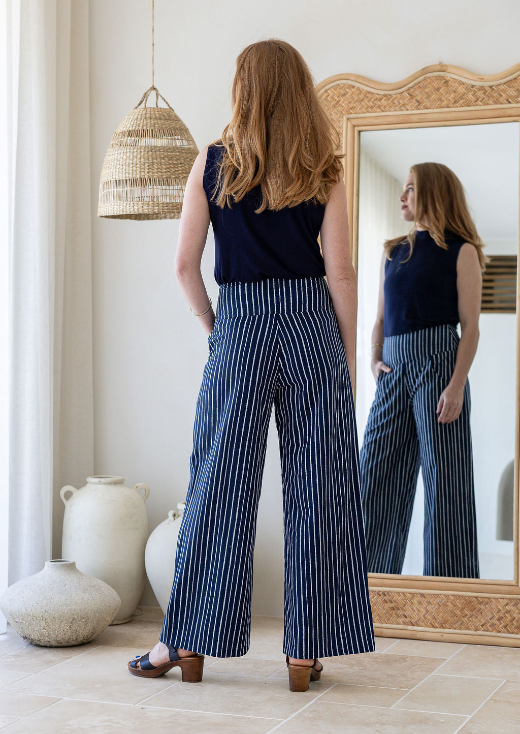 Model stands backwards with one hand in pocket in navy blue based white striped cotton pants with side zipper and wide leg fit