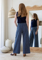 Model stands backwards with one hand in pocket in navy blue based white striped cotton pants with side zipper and wide leg fit