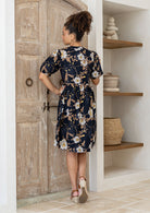 Woman stands backwards wearing a knee length relaxed fit 100% cotton dress standing in a room with wooden door and shelves.