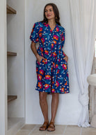 Woman wearing a knee length relaxed fit button through 100% cotton dress with elbow length sleeves and hand in pockets.