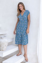 Woman stands in blue based floral print rayon midi length dress with buttons through the centre front and elastic through the waistband