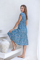Woman standing sideways in blue based floral midi length rayon button through dress with V-neckline and elasticated waistband