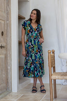 A woman modeling a short-sleeve midi dress with a vibrant floral print in teal, green, yellow, and orange on a dark navy background.