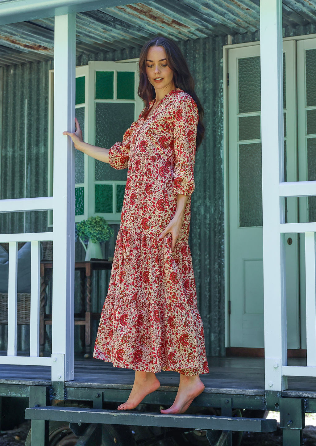 Woman stands side ways wearing a 100% cotton tiered maxi dress in cream based floral print with 3/4 sleeves, pockets and mandarin collar