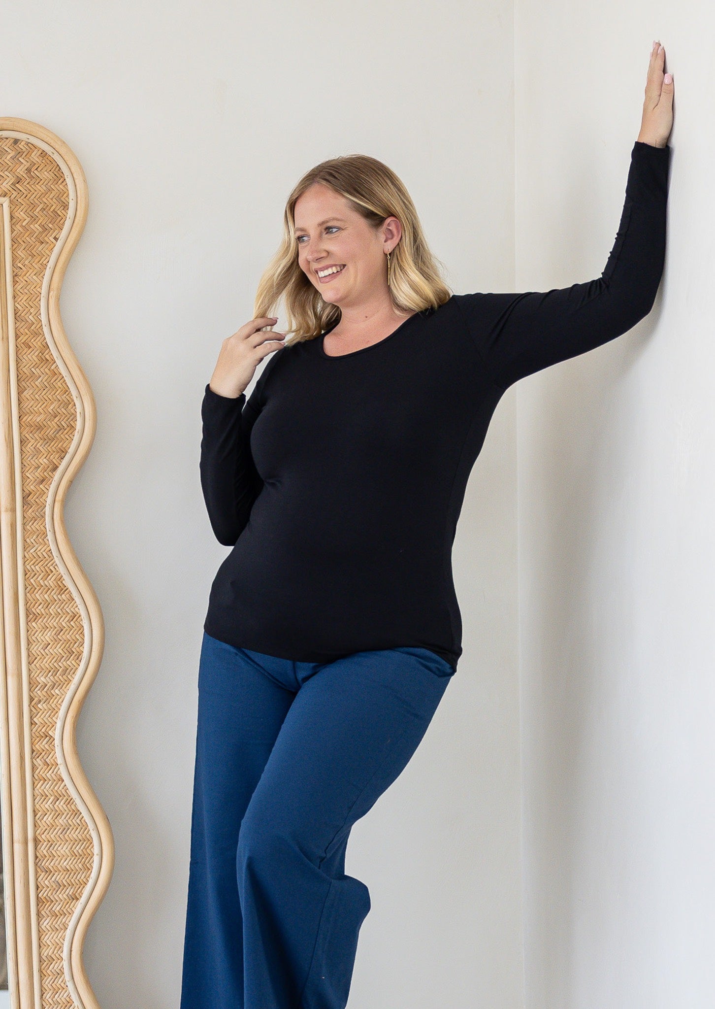 A woman with blonde hair wearing a black rayon jersey long-sleeve round neck top and blue pants, posing against a white wall