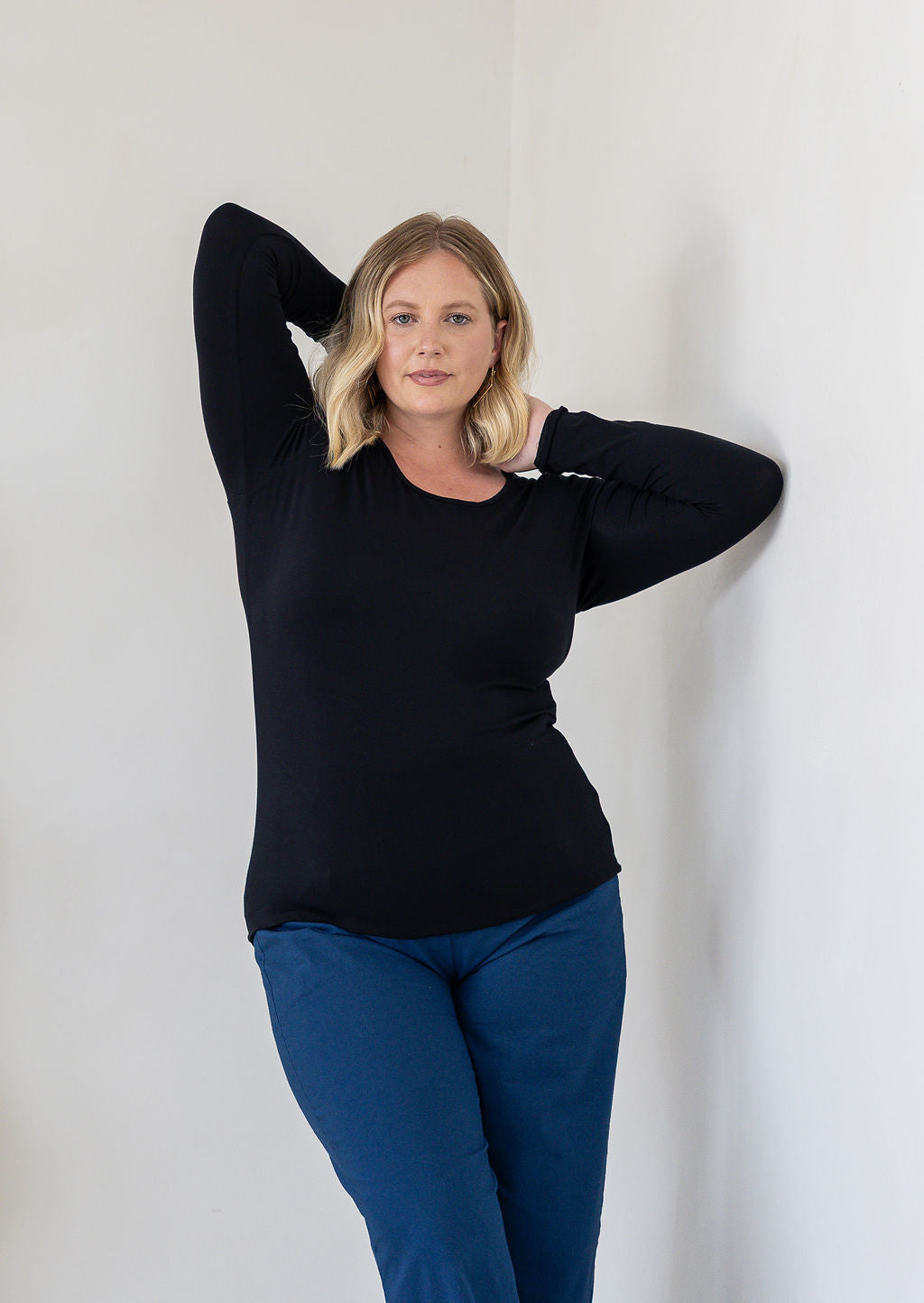 A woman with blonde hair posing in a black long-sleeve stretch rayon round neck top and blue pants against a plain white wall.
