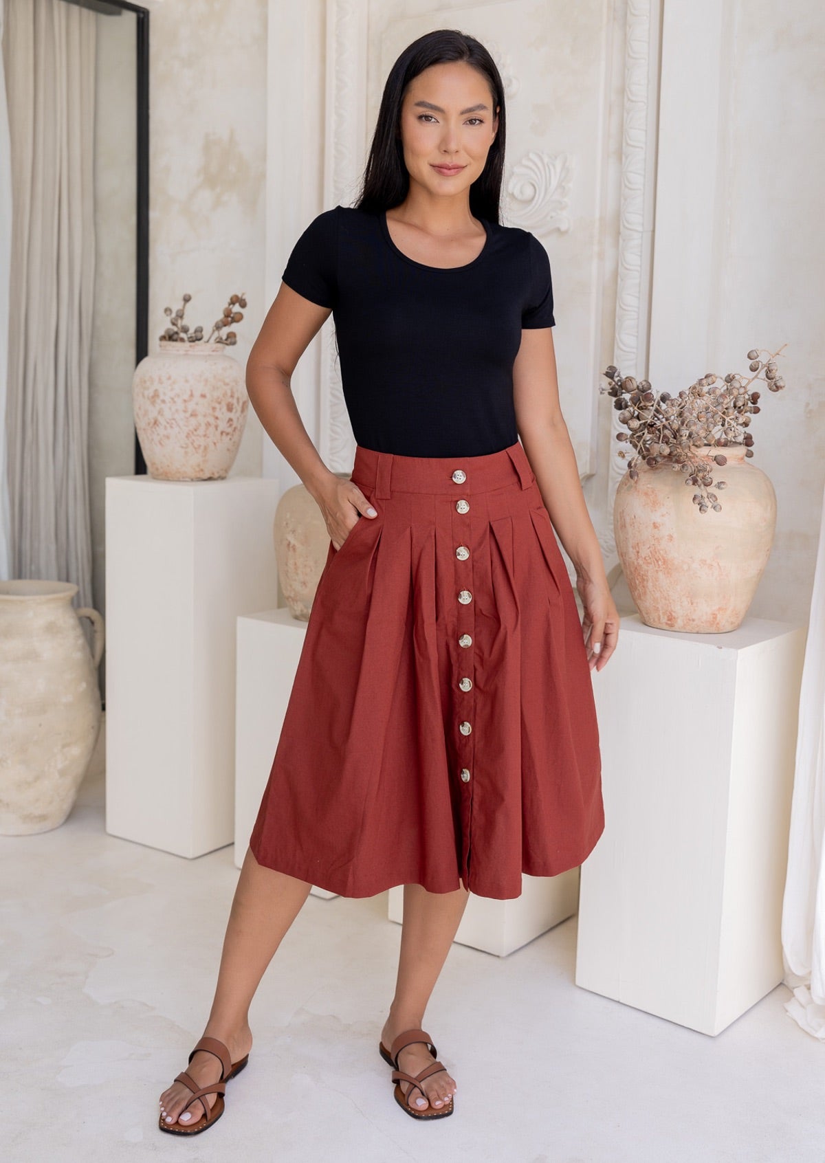 Woman wears rust coloured cotton A-line button through skirt with box pleats and belt loops, paired with a black t-shirt