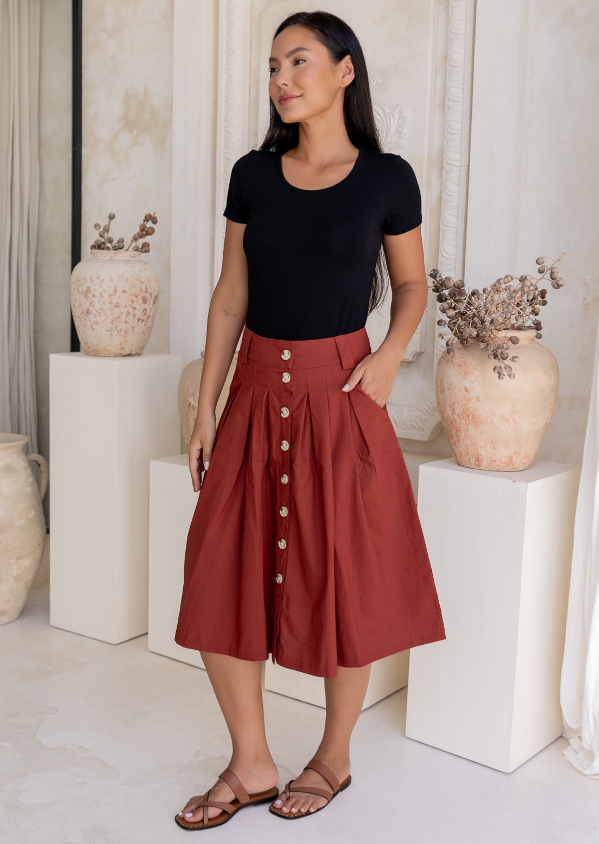 Woman wears rust coloured cotton A-line button through skirt with box pleats and belt loops, with hand in pocket