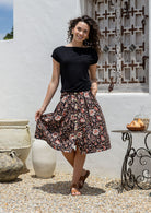 Woman stands wearing a basic black top and black based floral print, button through cotton skirt with box pleats and belt loops 