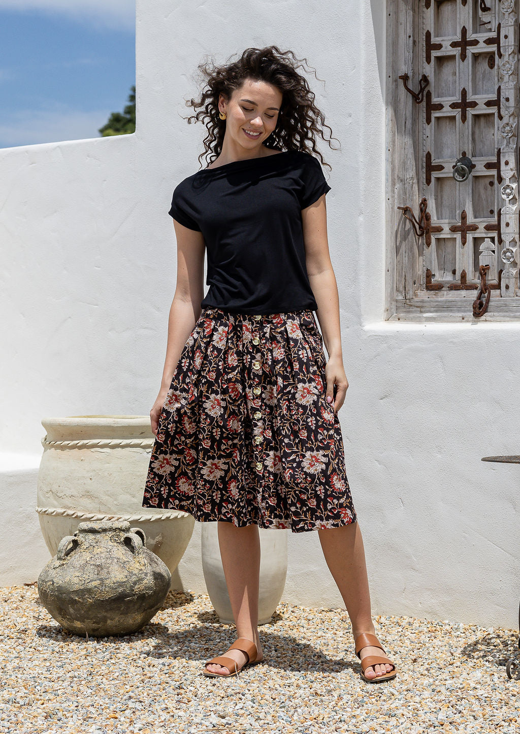 Woman wearing a knee length, box pleated cotton skirt with button front, belt loops and pockets in black based Indian traditional print